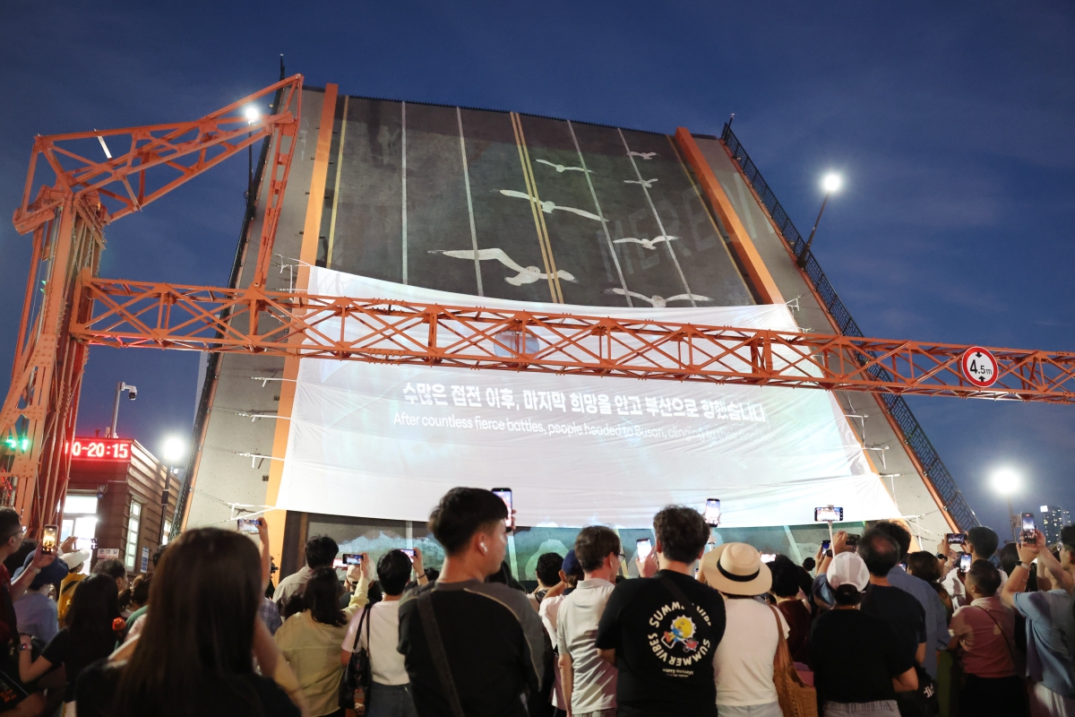 ２５日、釜山・中（チュン）区の影島（ヨンド）大橋で「２０２６避難首都 釜山 国家遺産ナイトツアー」が開催された。写真は、市民らが橋桁が開くパフォーマンスとともに、１０２３日間に及ぶ避難首都・釜山の歴史を描いた映像を鑑賞している様子。１９３２年に着工した影島大橋は、釜山・中区と影島区を結ぶ韓国初の連陸橋（陸地と島を繋ぐ橋）だ。韓国戦争当時、人々の出会いと別れの舞台となった避難首都・釜山を代表するシンボルである。