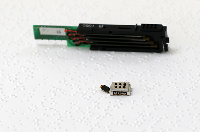 ドットが独自開発した超小型点字セル(下は能動アクチュエータ)と従来の点字端末に使用された中核部品セルが点字書籍の上に置かれている。この超小型の点字セルはこれからドットのあらゆる製品に組み込まれ、これまで点字の使用や補助機器のコストのため様々な不便を強いられた視覚障害者の生活を改善する