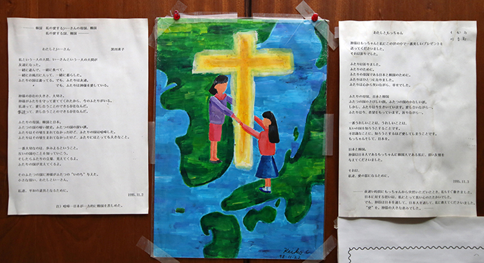 週報を手に持った吉田牧師(上)。週報には韓国と日本との間に十字架が置かれた絵が描かれている。両国が十字架を介して手を繋いで行こうというメッセージが込められている。ある青少年信徒が週報を見て描いた絵が教会の入り口に貼り付けられている(下) 